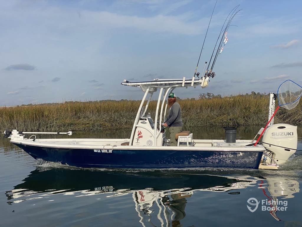 A center console fishing boat being navigated by a captain on the backcountry waters of Savannah, GA, with trolling rods perched above the helm and a net on the back of the boat