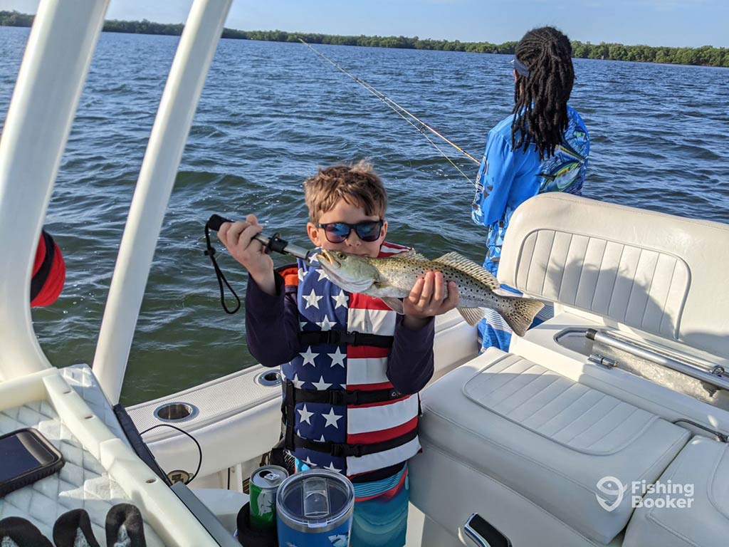 A young child in an American flag life vest holds a Speckled Trout up in front of his face, while a girl fishes off the side of the boat behind him on a sunny day