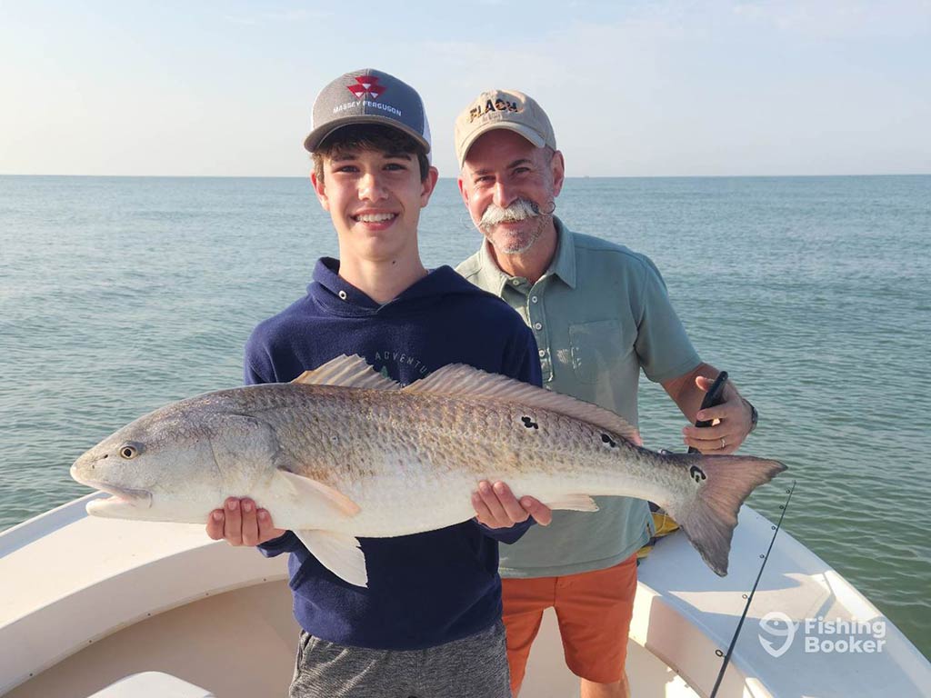 A teenage boy holding a Redfish aboard a fishing boat in Apalachicola on a sunny day, while an elderly man stands bihind him smiling towards the camera