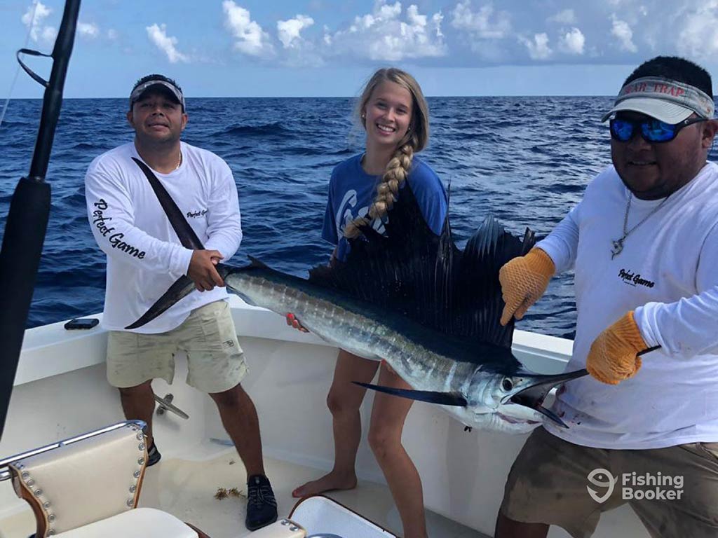 A woman stands between two men aboard a fishing boat in Puerto Morelos, as they hold a large Sailfish towards the camera, with the water behind them on a sunny day