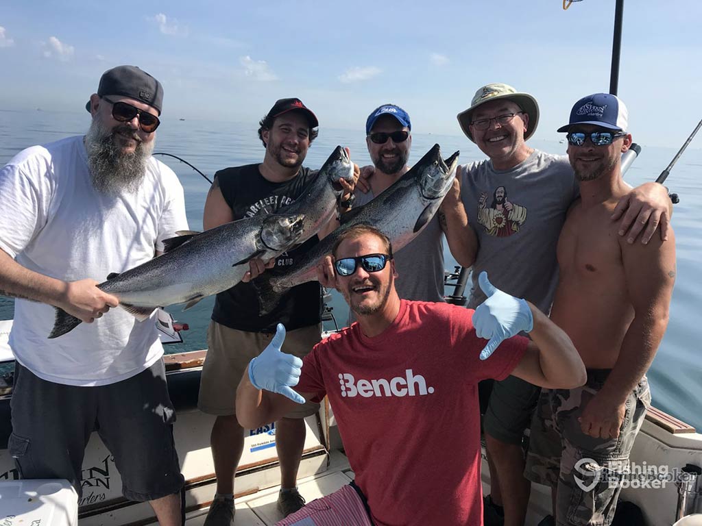 A group of male anglers on a fishing charter in Canada, the majority of which are holding a Salmon, while one man in a red shirt crouches in front of them with his thumbs up