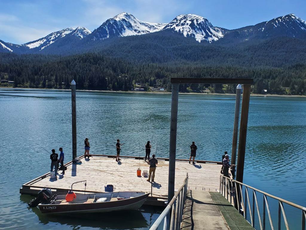 A view from a hill of a small fishing dock in the Gastineau Channel, full of anglers on a sunny day with snow-capped mountains in the distance