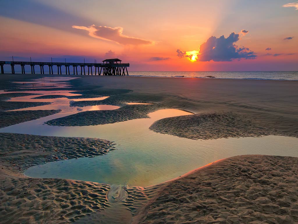 A view across the beach towards a fishing pier in Tybee Island, with puddles in the sand visible in the foreground and the sun setting in the distance