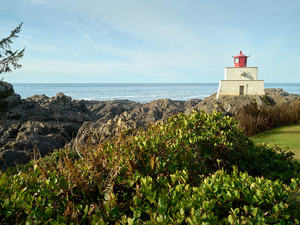 A view across a green landscape towards a short lighthouse with a red top, with the ocean in the distance on a sunny day in Ucluelet