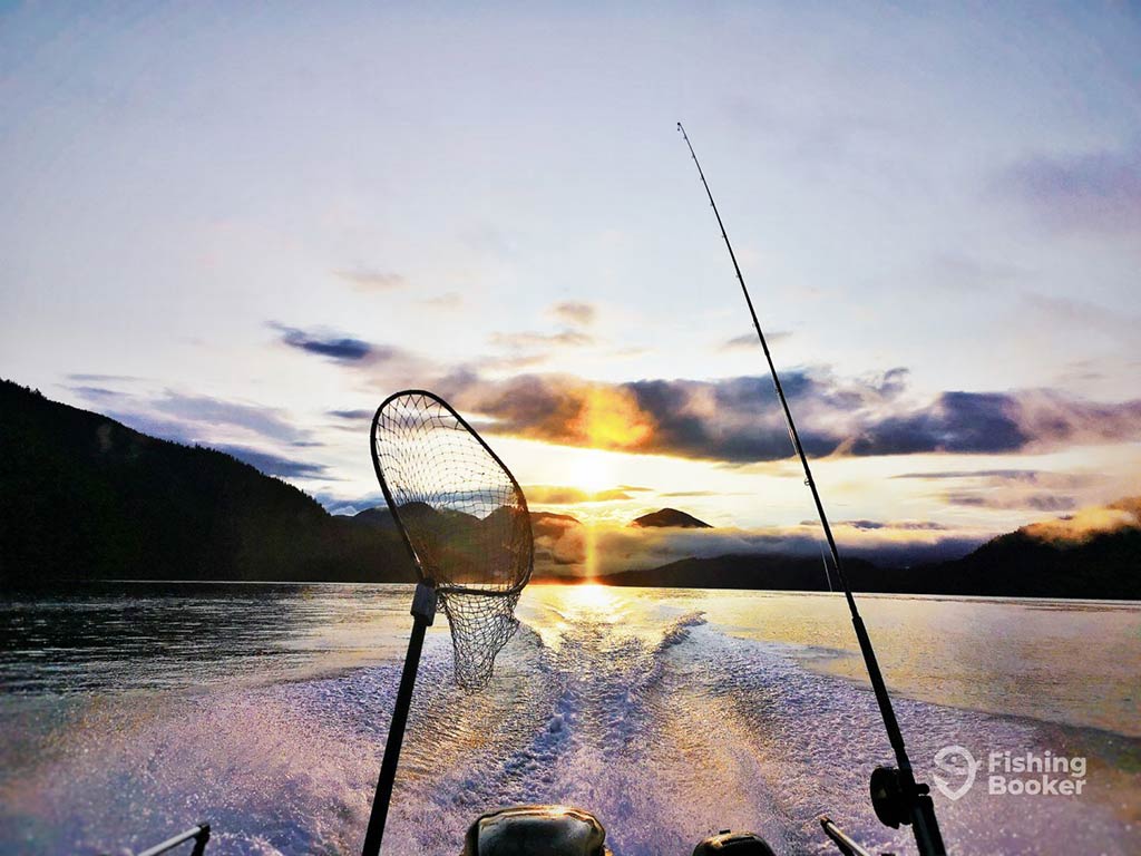 A view towards the back of a fishing boat with a lone trolling rod and fishing net perched towards the water and the sun setting in the distance in Ucluelet