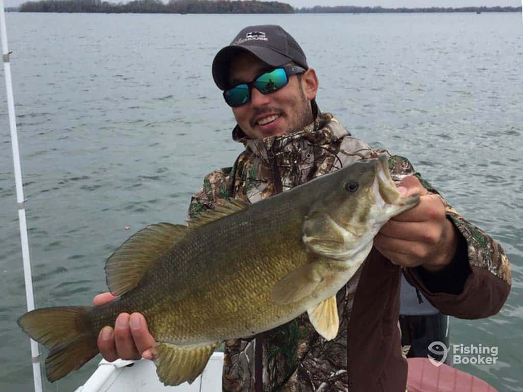 An angler leans back while holding a Smallmouth Bass, caught while fishing near St. Catharines in Ontario on a cloudy day, with the water behind him