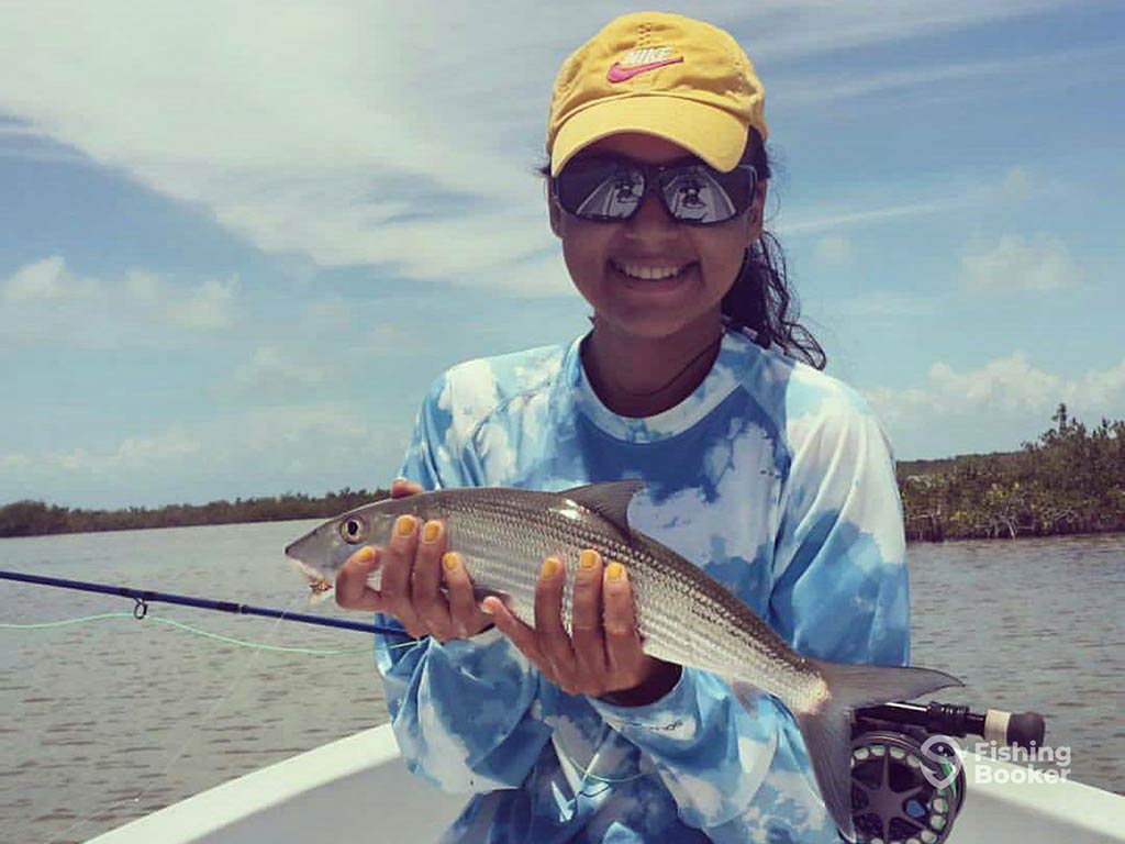 A woman in sunglasses and a yellow baseball cap holds her Bonefish, along with a fly fishing rod, aboard a fishing boat in the flats of San Pedro, Belize