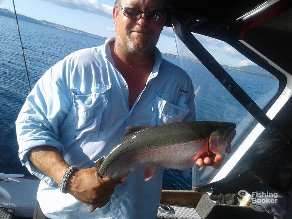 An angled photo of a man standing on a charter boat and holding a Trout, with a lake's waters behind him on a clear day