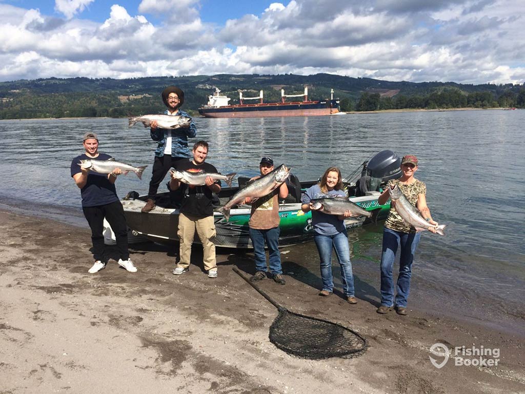 A group of anglers on the shore of the Columbia River holding up a Salmon each on a cloudy day with a boat behind them and a big ship in the distance on the opposing shore