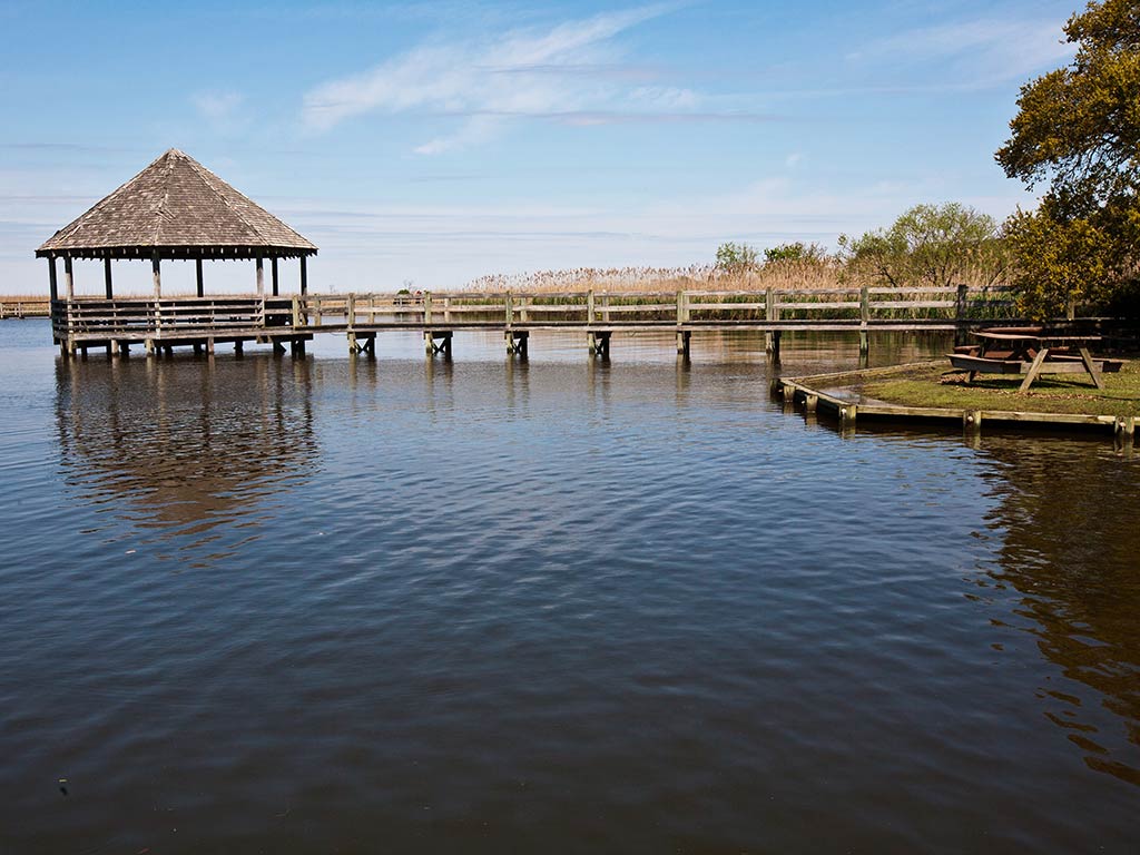 A view across the calm waters of Currituck Sound, NC, towards a small, wooden fishing pier on a sunny day