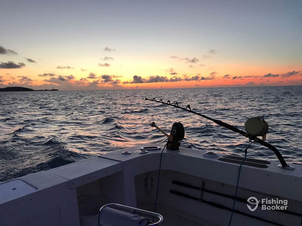 A view of a heavy-duty fishing rod with electric reel hanging off the side of a boat in Bermuda at sunset, with the water and sun setting in the distance