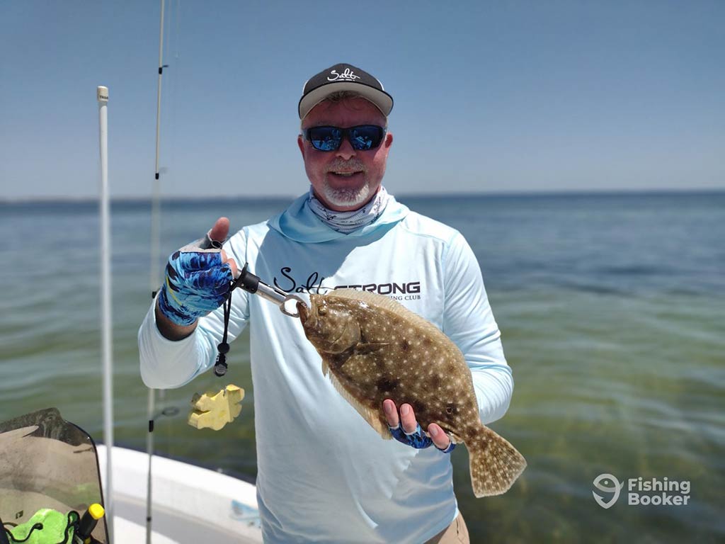 An angler in a baseball cap and sunglasses holds a Flounder up to the camera while standing on a fishing boat, with the murky backcountry waters behind him on a clear day