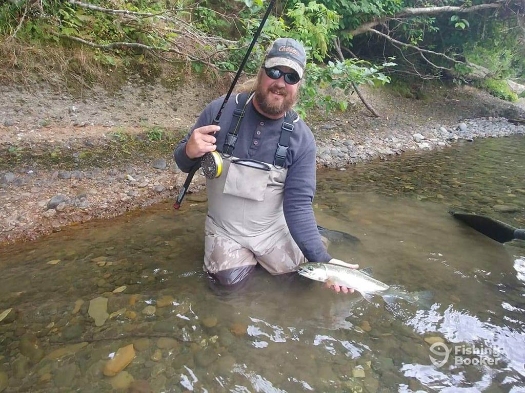 An angler up to his thighs in a river, holding a fly fishing rod in one hand and a small Salmon, submerged in the water in the other with the bank behind him on a grey day