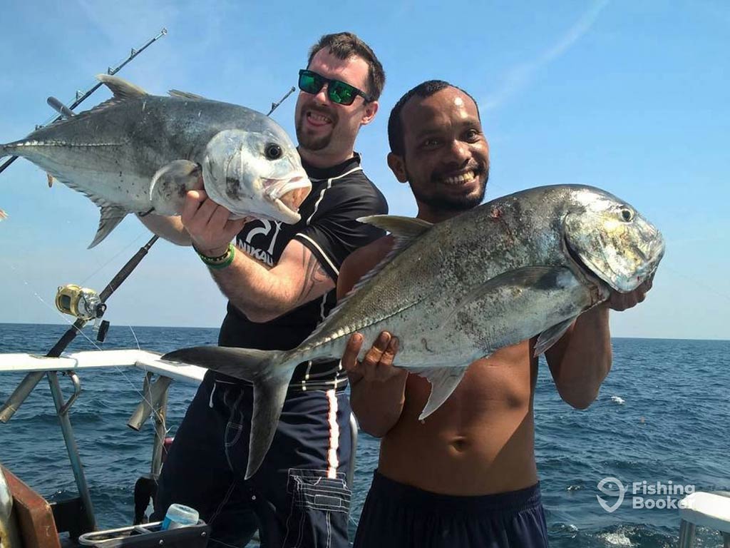 Two anglers present a Giant Trevally each to the camera aboard a fishing charter in Phuket, with the water and clear blue skies visible behind them