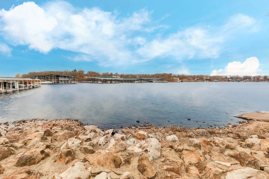 A view of the rocky shores of the Grand Lake O' the Cherokees in Oklahoma, with calm waters and slightly cloudy skies, along with docked boats visible across the lake.