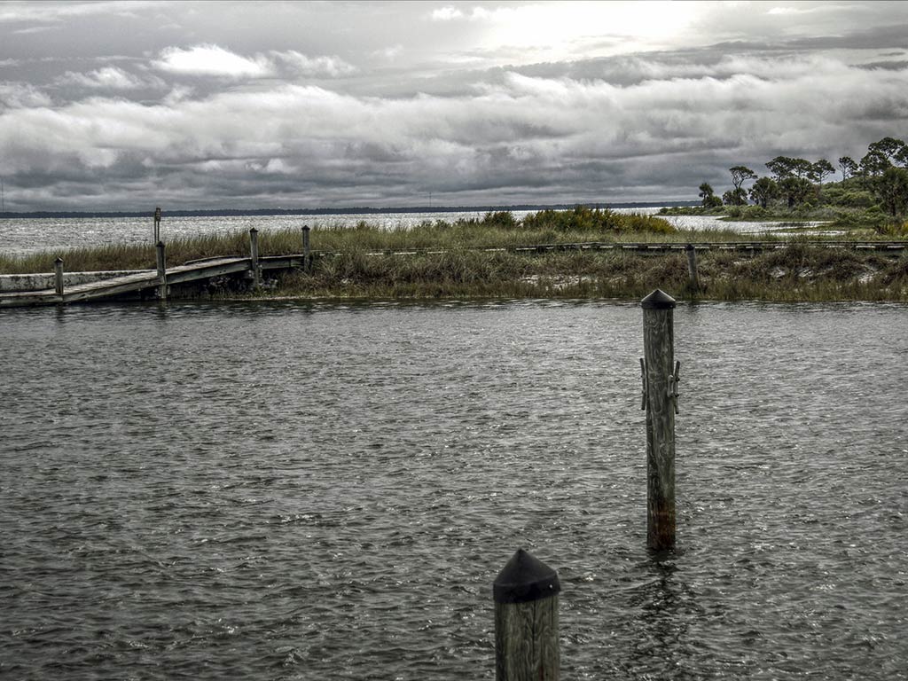 A view across the water towards a small, wooden fishing pier or boat dock in the inshore waters near Cape San Blas on a cloudy day