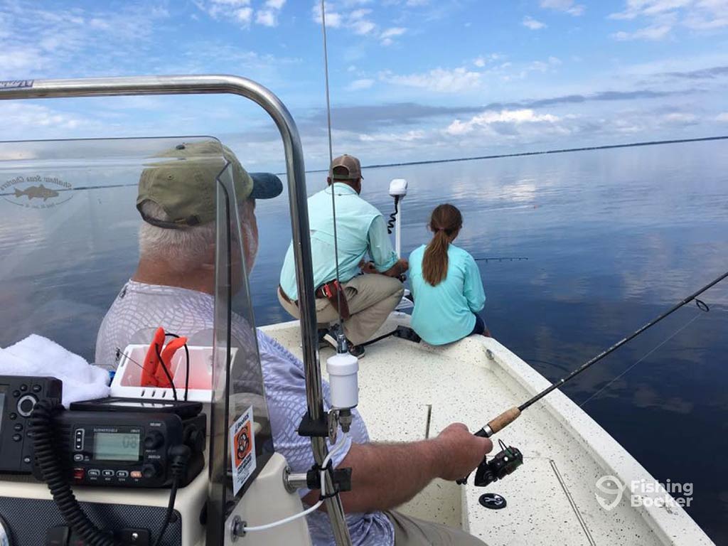 A view from behind of a middle-aged angler fishing over the side of a fishing boat in calm, inshore waters, with a man and a young girl also fishing over the front of the boat on a day with sunny intervals