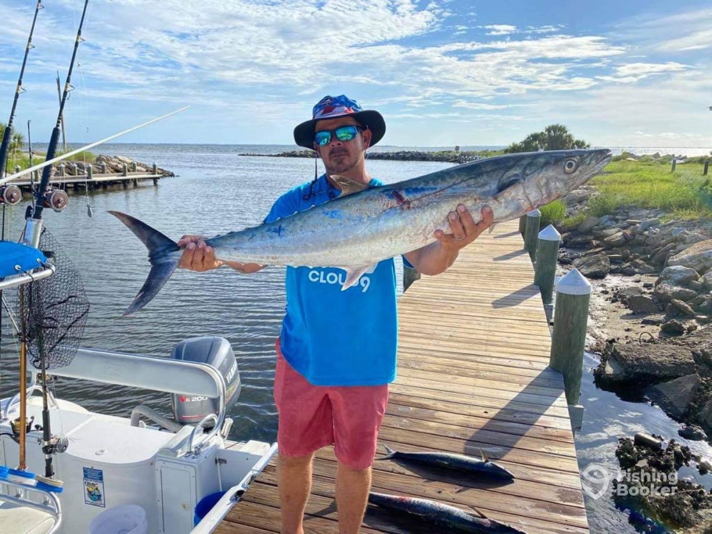 An angler wearing sunglasses and a hat stands on a boat ramp in Cape San Blas, holding a large Kingfish caught while fishing the reefs of the Gulf of Mexico on a sunny day, with the water visible behind him, along with some backcountry mangroves