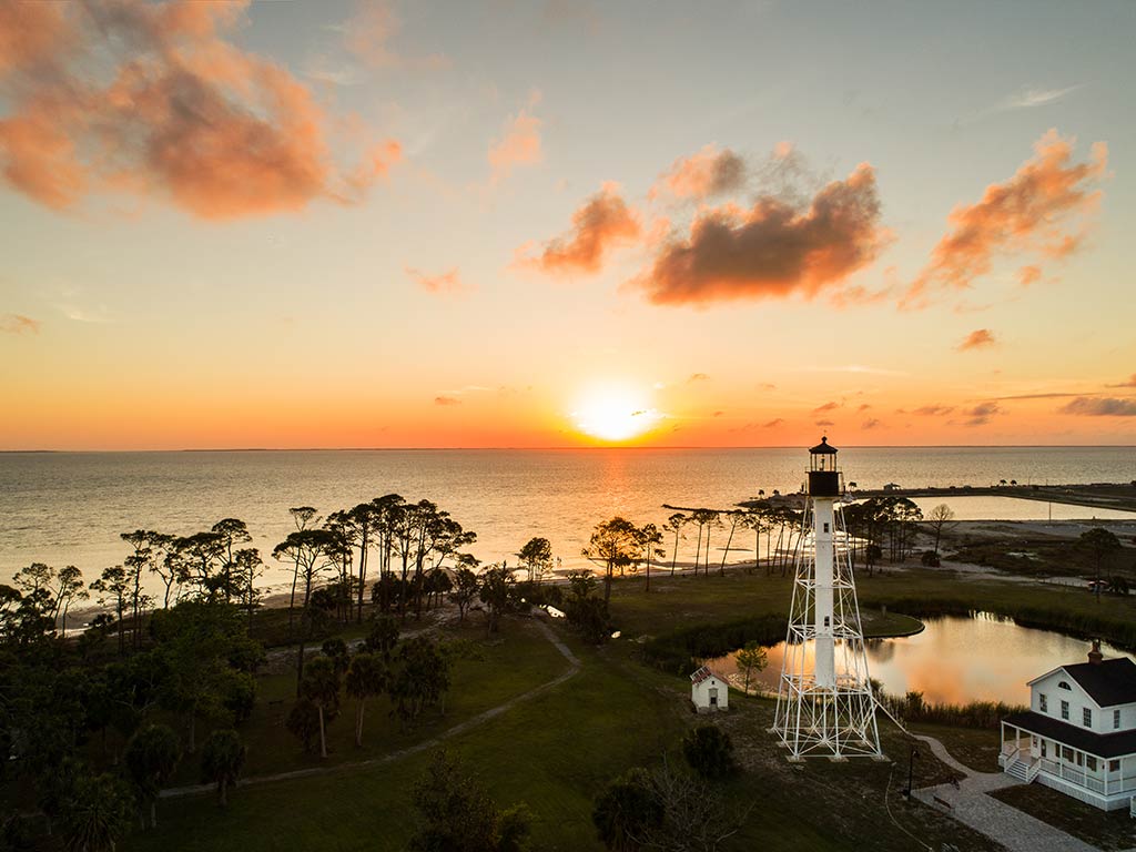 A view towards a lighthouse at Cape San Blas at sunset, with the sun setting in the distance over the Gulf of Mexico and some greenery visible in the foreground