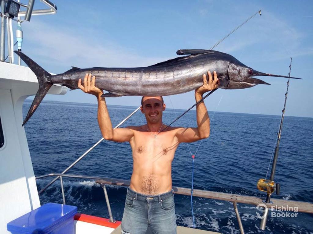 A shirtless angler holds a large Marlin above his head aboard a fishing charter in Phuket, with the water behind him on a clear day