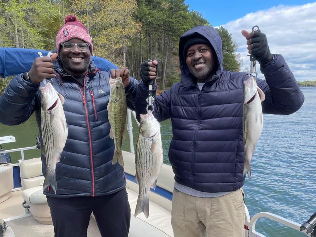 Two anglers in winter gear, standing on a fishing boat and holding a mixture of different Bass fish in their hands on a day with sunny intervals