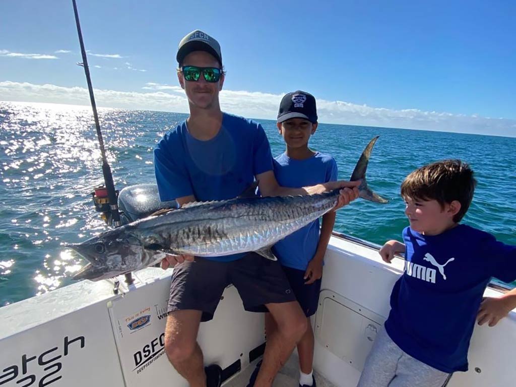 A teenager holds a large Narrow-Barred Spanish Mackerel with two younger boys looking on, at the corner of a fishing boat with the water behind them on a sunny day