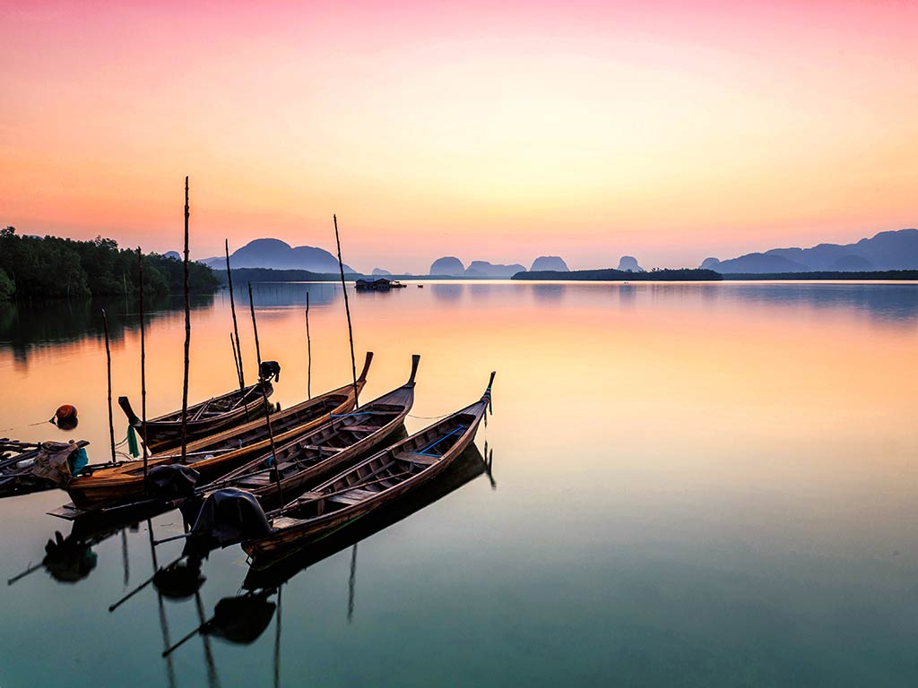 A view from the shore across three traditional, wooden Thai fishing boats towards the sea, with the sun setting in the distance, creating an orange-pink hue on the water and in the sky