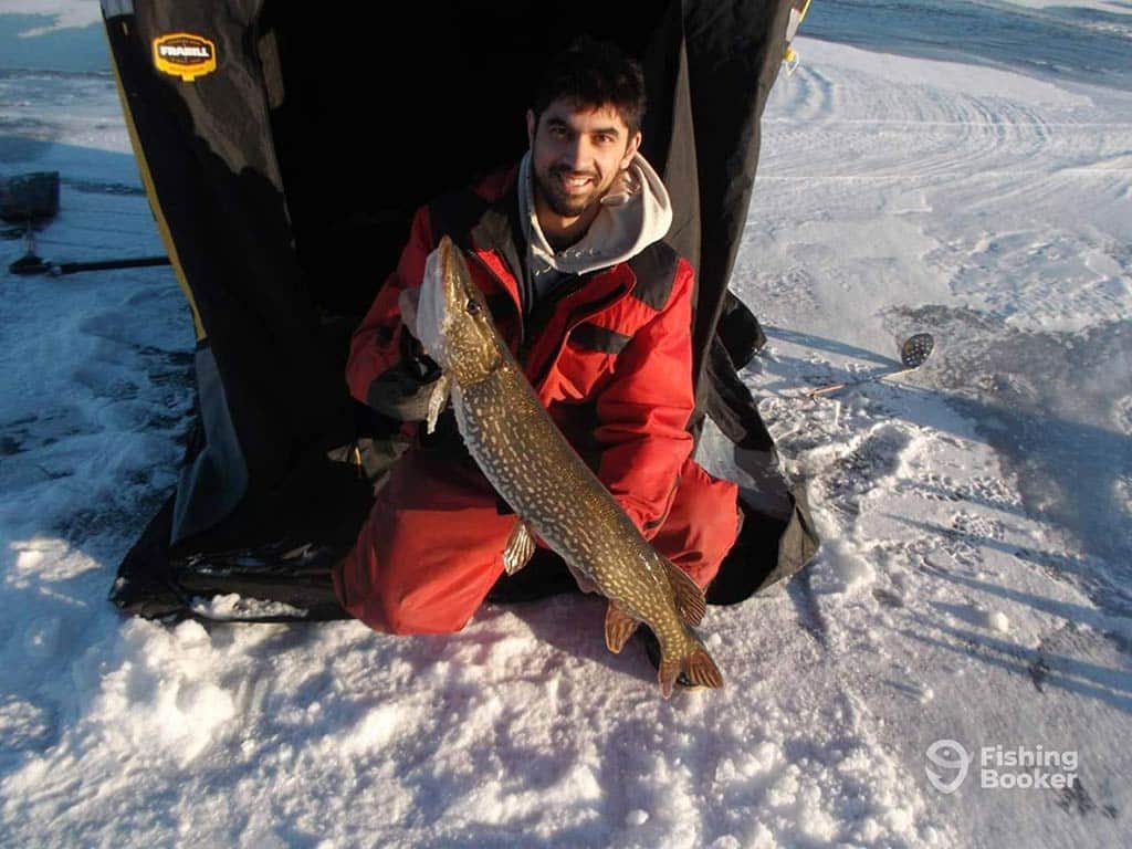 An ice angler in red overalls holding up a Pike, caught while ice fishing in Ontario on a sunny day, with his ice hut behind him