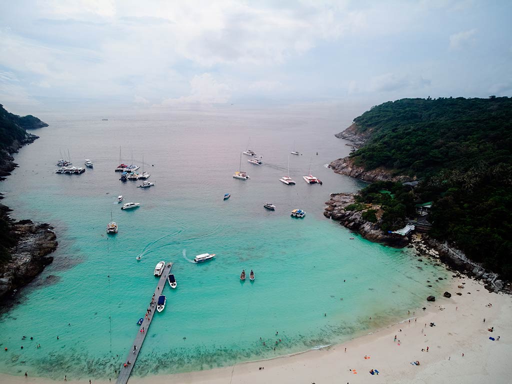 An aerial view of a small marina in Racha Noi, Phuket on a cloudy day with turquoise waters visible in the foreground and greyer waters in the distance