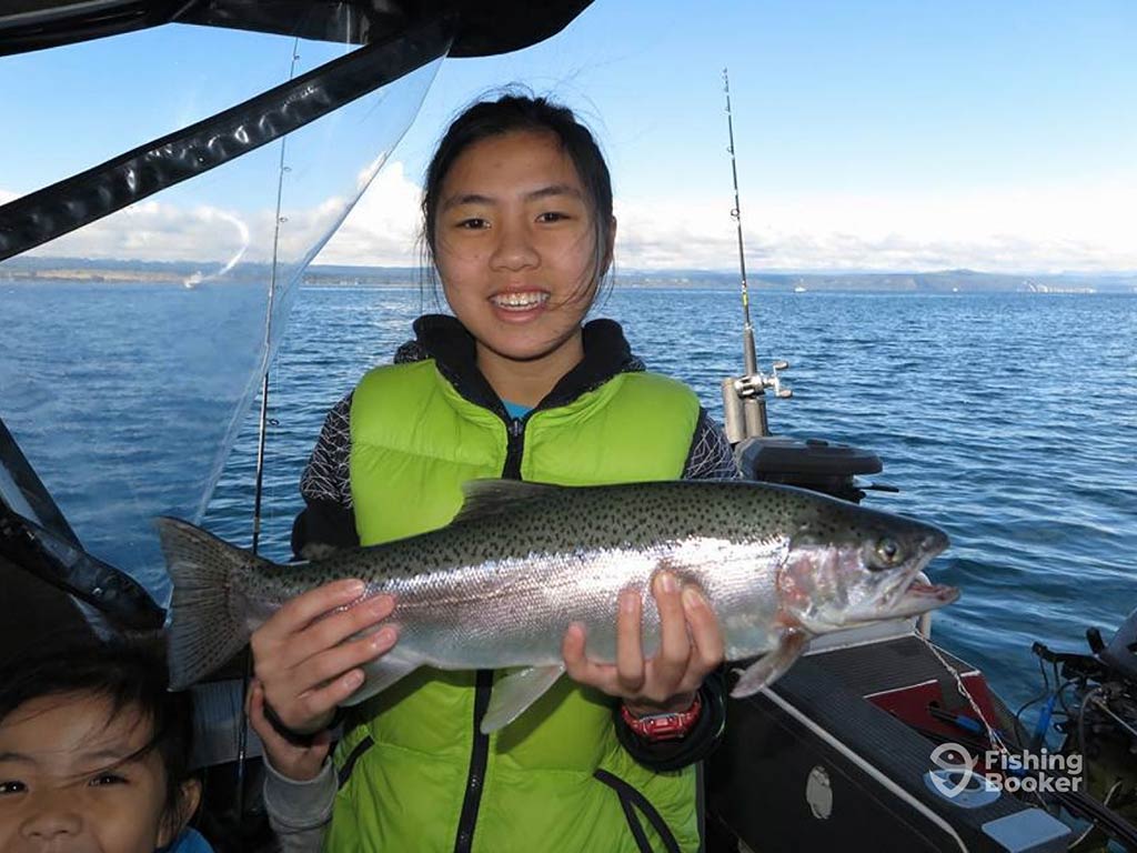 A young girl in a fluorescent green life vest, holding a shiny Rainbow Trout caught while fishing in Taupō, with the water behind her