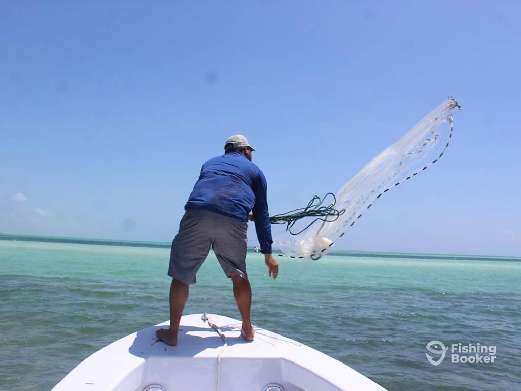 A rearview image of an angler casting a net along the reefs from the front of a fishing boat in turquoise waters on a clear day