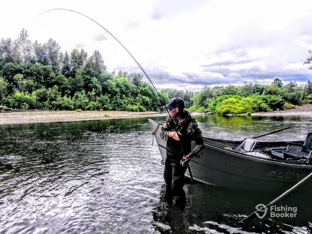 A man wading in a river next to a fishing boat in Washington, with his rod bent and cloudy skies and shorelines visible around him