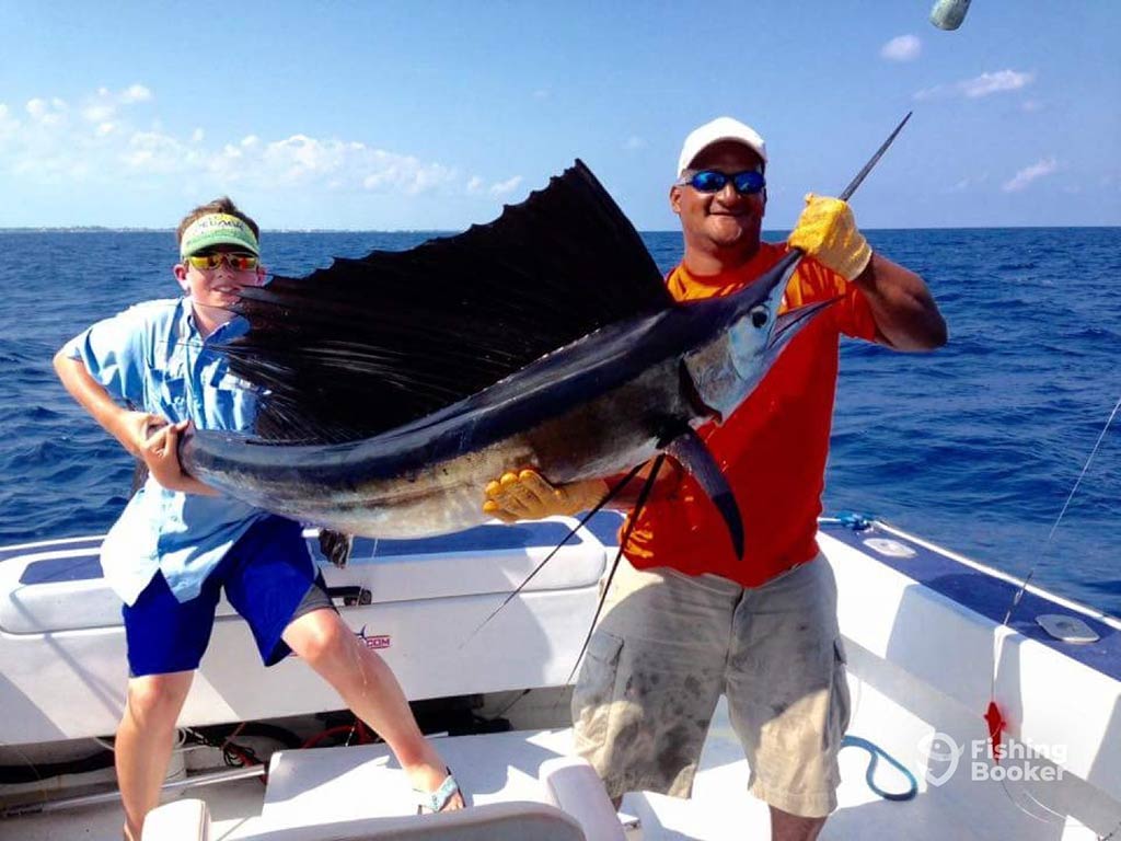 A youth and a man hold a large Sailfish from either end aboard a sportfishing boat offshore from San Pedro, Belize on a sunny day, with the water behind them