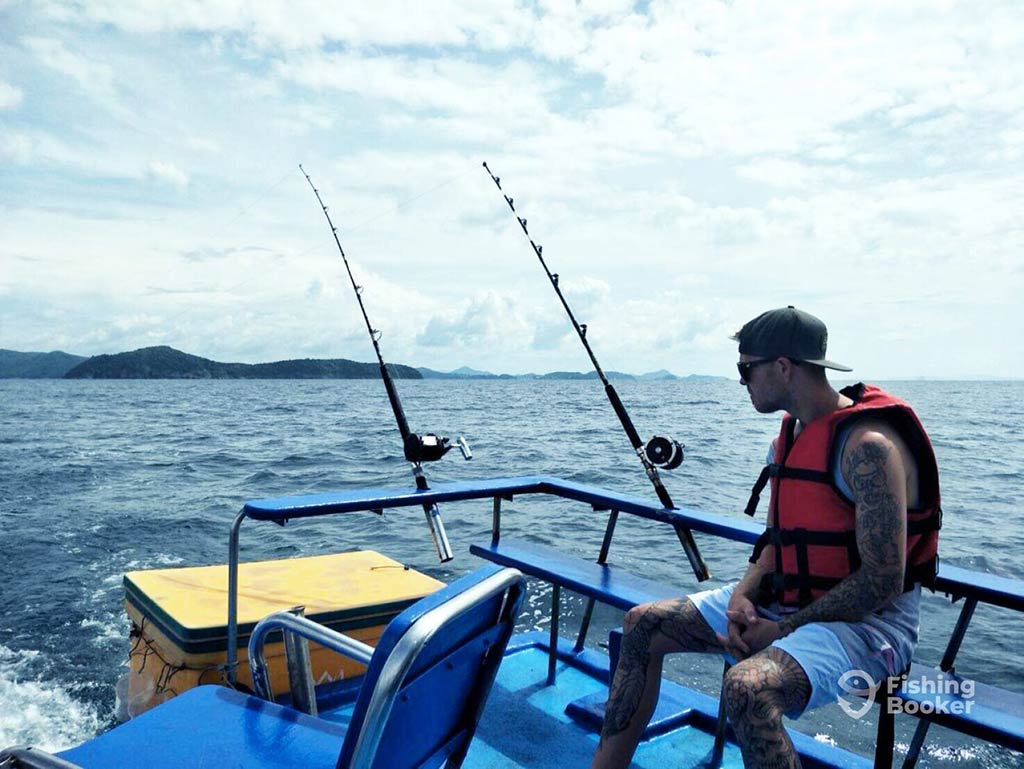 An angler in a life vest sits looking out of the back of a fishing boat in Phuket, with two trolling rods set up and some land visible in the distance on a cloudy day