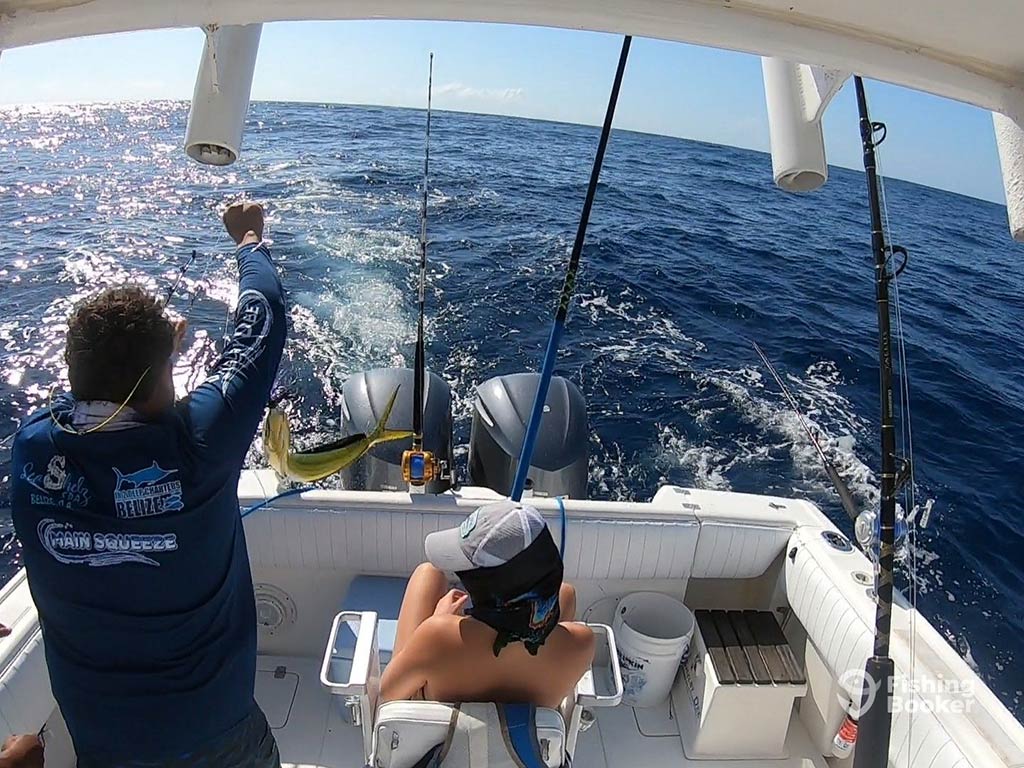 A view out of the back of a fishing boat in San Pedro, Belize, with two fishing rods trolling and one angler trying to hook a Mahi Mahi aboard, while a woman sits in a fighting chair