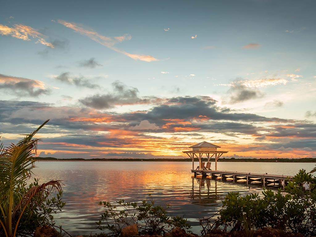 A view out towards a small, wooden fishing pier at sunset in San Padro, Belize, with the sun behind the clouds in the distance