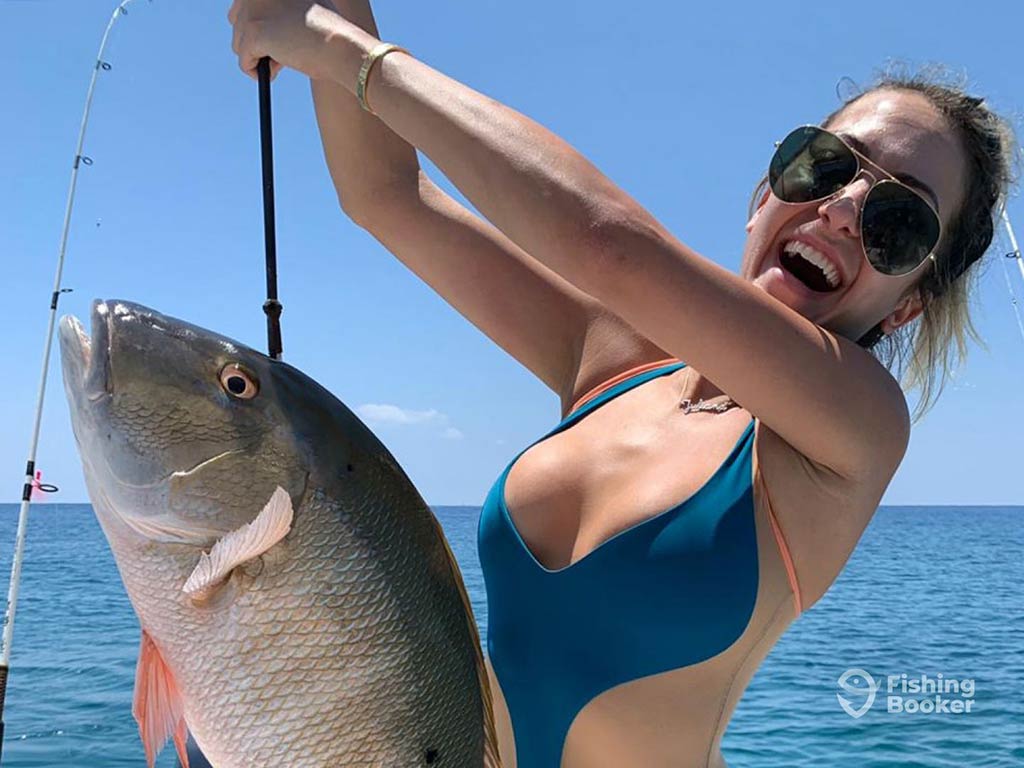 A closeup of a woman in a one-piece swimsuit and sunglasses holding a Snapper by a fishing pole with the water behind her on a sunny day in Belize