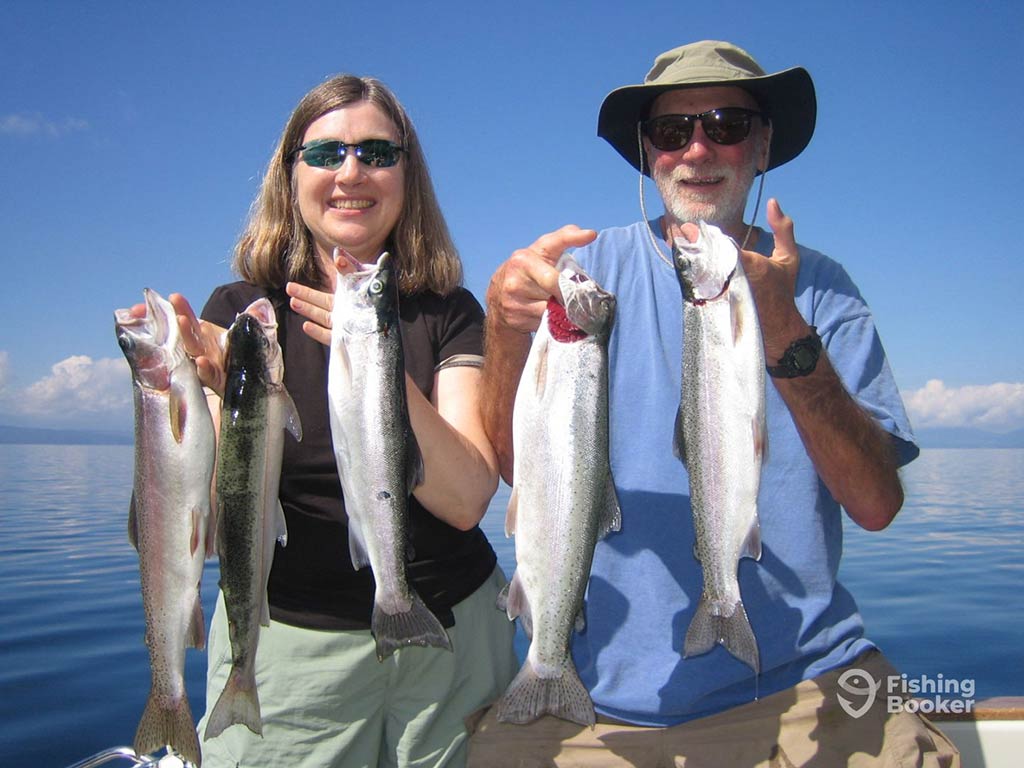 A man and a woman holding up their freshly-caught Trout aboard a fishing boat in Taupō on a sunny day, with the water behind them and blue skies above them