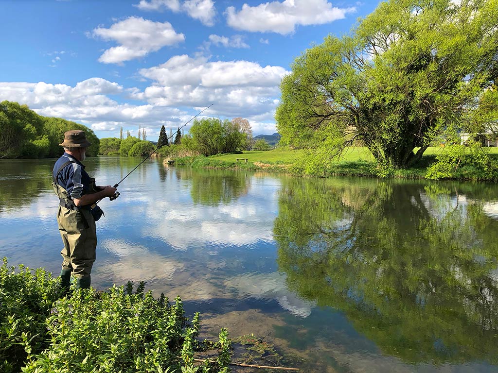 A man in full wading gear standing on the shore of a river near Taupō while fly fishing, with the reflection of the sky and the surrounding trees visible on the water