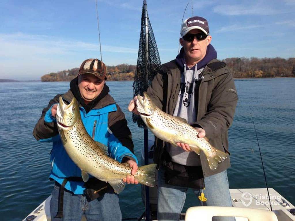 Two anglers proudly present their Trout catches on a fishing charter near St. Catharines, Ontario, with the water and some land visible in the distance behind them on a clear day