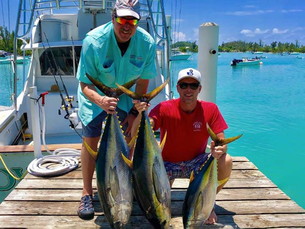 Two men on a dock in Bermuda, holding three Tuna between them, with a large offshore sportfishing vessel and crystal clear blue waters behind them on a sunny day