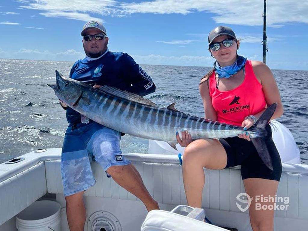 A man and a woman lean on the back of a fishing boat, as they struggle to hold a large Wahoo caught while deep sea fishing in San Pedro, Belize on a clear day