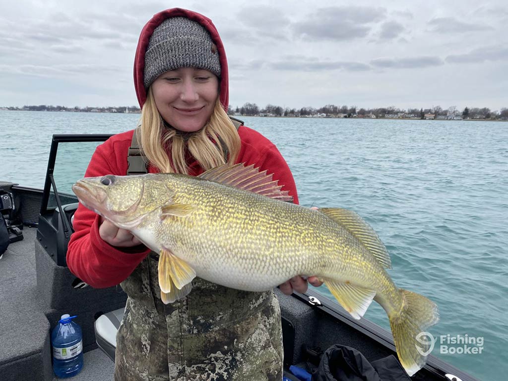 A blonde, female angler in a woolly hat and hood, holding a Walleye, while looking down at it, standing on a fishing charter in the Great Lakes region in Ontario on a cloudy day