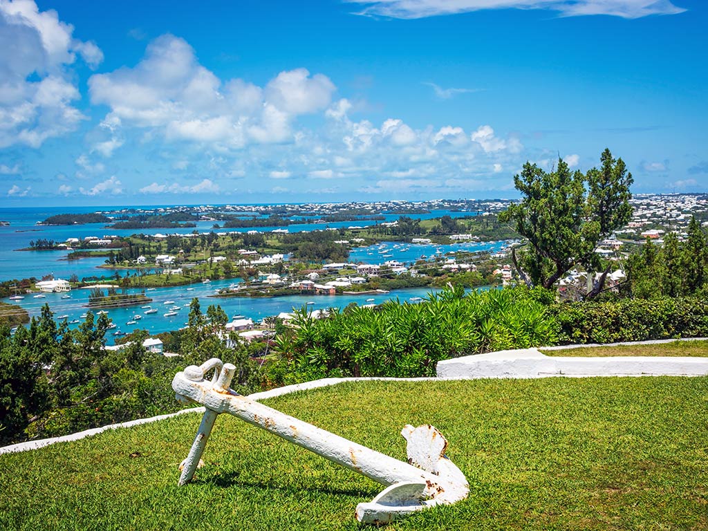 A view from a hill towards a bay in Bermuda, with trees and houses in the distance, and an anchor in the foreground on the hill on a sunny day