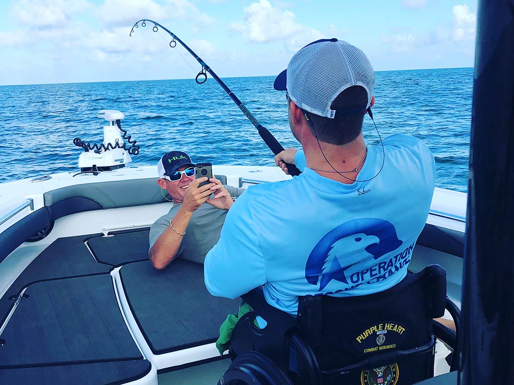 A view from behind of an angler in a wheelchair bottom fishing off the front of a boat, with another man in front of him taking a photo and smiling