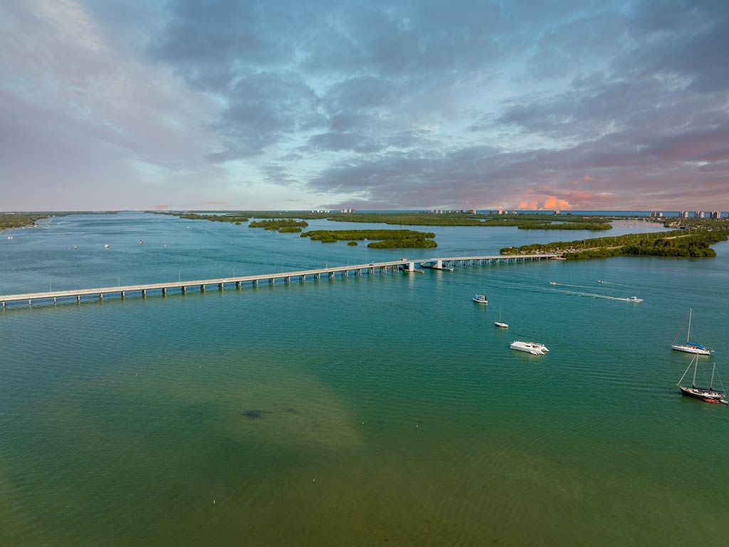 An aerial view of a bridge across the Indian River Lagoon, with some mangroves visible in the distance at sunset on a relatively clear day, with a few clouds visible in the sky