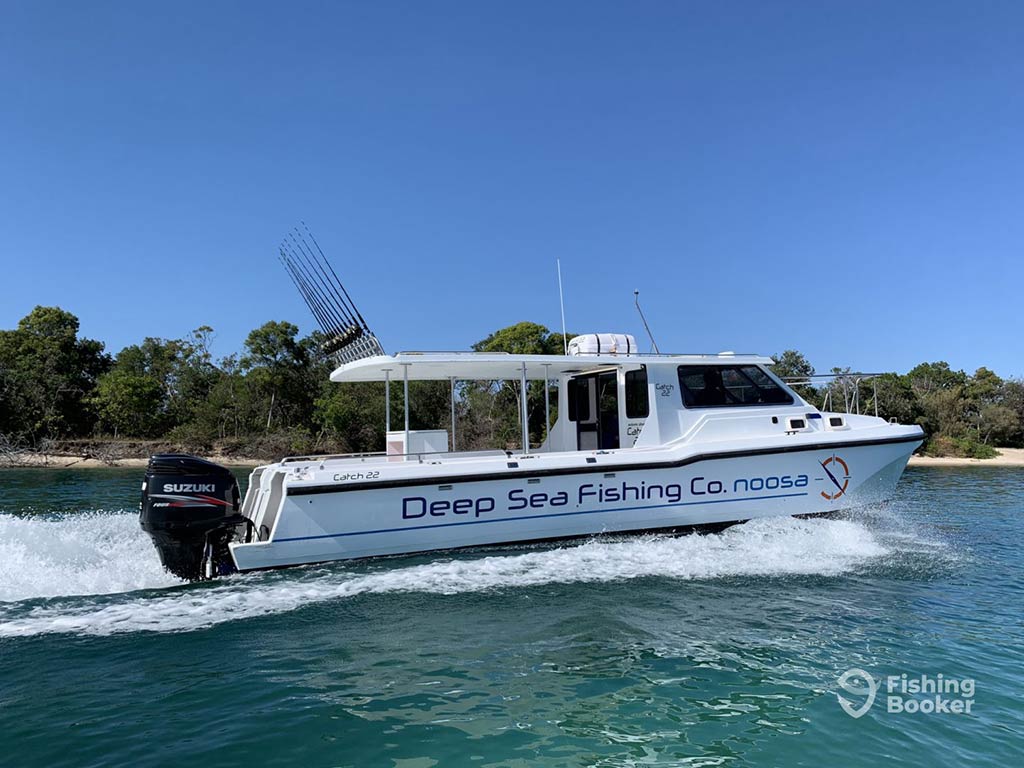 A view across the water towards a fishing charter with the wheelhouse at the front of the boat and trolling rods hanging above the back of the boat on clear blue waters on a sunny day