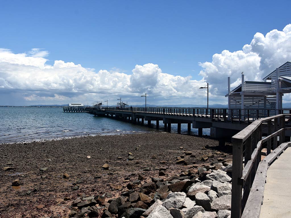 A view from a boardwalk towards the sea, with a fishing pier sticking out into the waters of Port Moreton on a sunny day