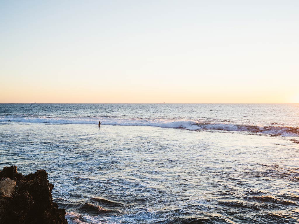 A view from shore towards some incoming waves in Scarborough, QLD, where a lone angler is knee-deep in the water casting a line at sunset