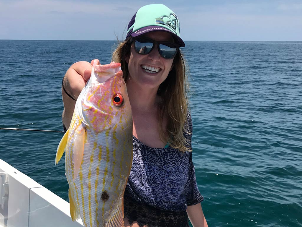 A woman in a baseball cap, holding up a small Snapper to the camera aboard a fishing boat, with the open water behind her on a clear day in Florida